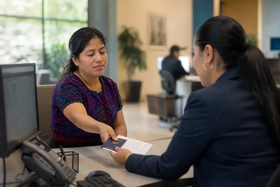 Mujer entrega su pasaporte y una factura a una empleada de una cooperativa de crédito